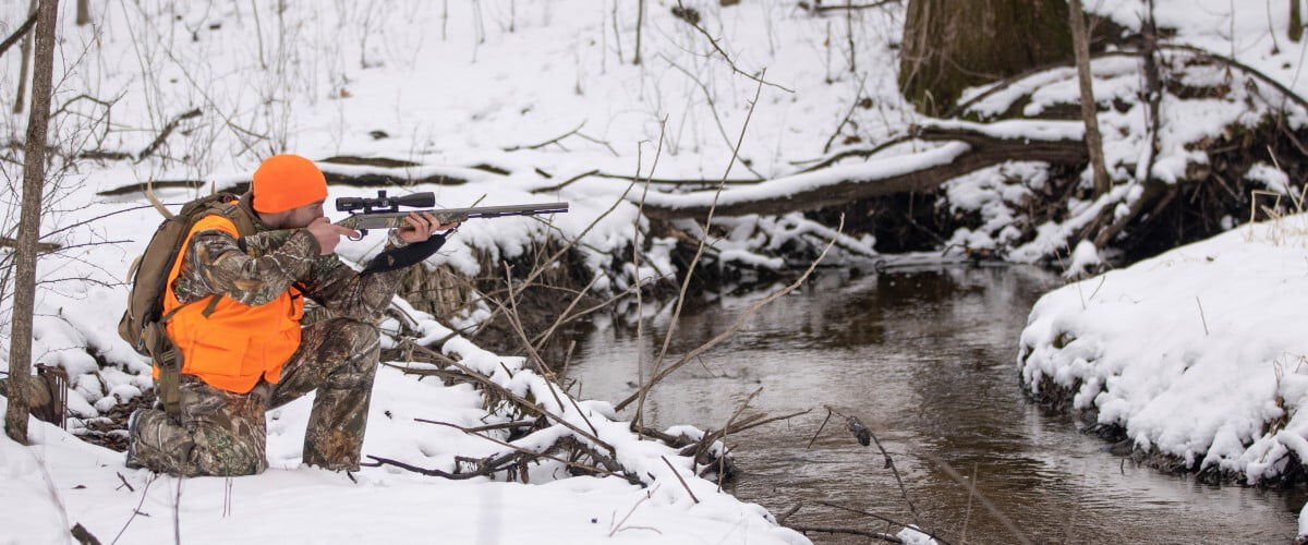 hunter kneeling in the snow while aiming a rifle