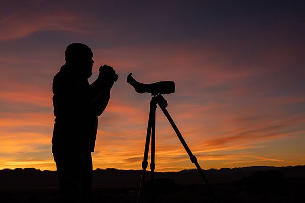 hunter holding binoculars next to a spotting scope during a sunrise