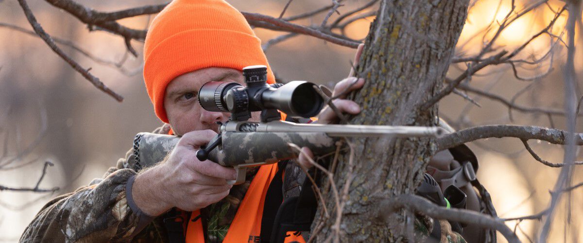 hunter looking down the scope of a rifle while resting the rifle on a tree