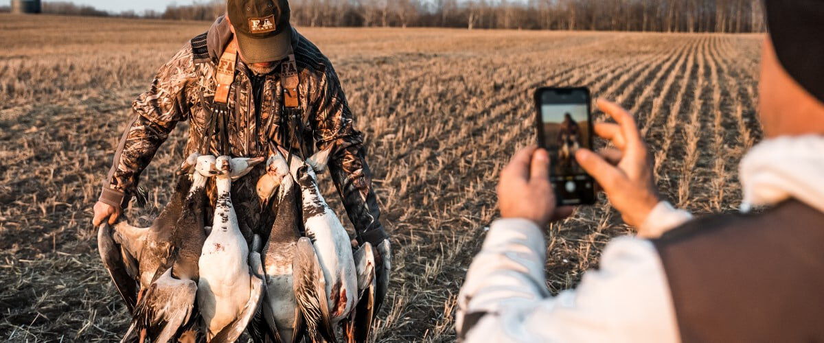 person taking a picutre on their phone of a hunter with a bunch of dead ducks draped around their nexk