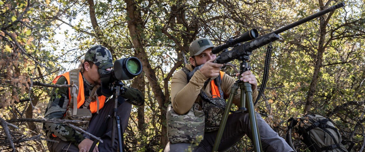 one hunter looking down the scope of a rifle on a tripod and another hunter looking through a rangefinder