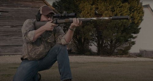 Jim Gilliland kneeling while looking down the scope of a rifle