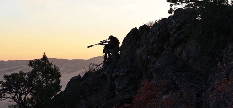hunting aiming rifle while sitting on a rocky hill