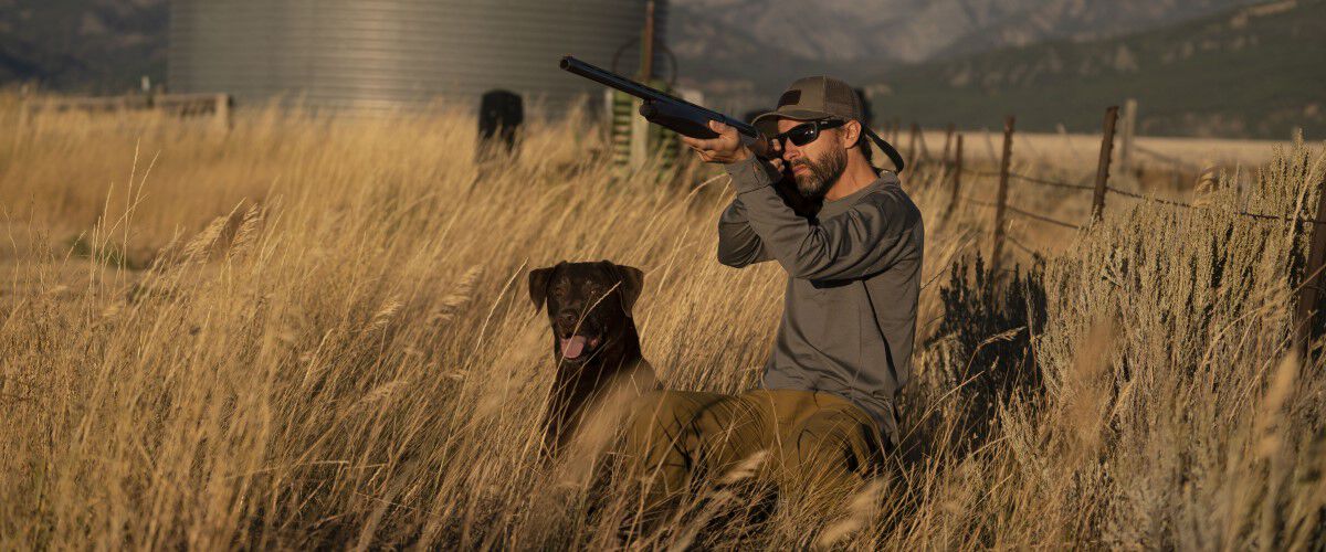 hunting dog sitting next to hunter who is aiming shotgun