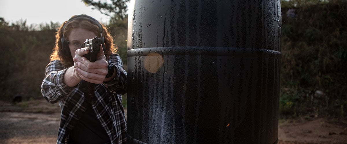 lady aiming handgun around a barrel outside