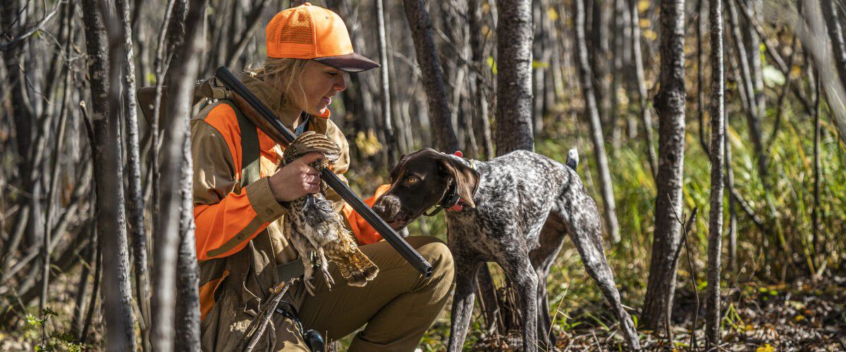 female hunter kneeling down next to a germain shorthair while holding a dead bird