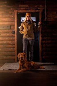 female wearing the Cirque jacket and standing in a doorway with a shotgun over her shoulder and a dog at her feet