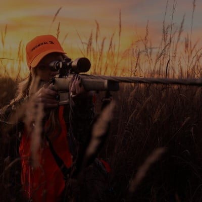 hunter in tall grass looking down a rifle scope