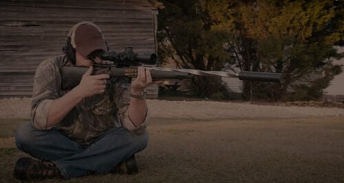 Jim Gilliland sitting on the ground and looking down a rifle scope