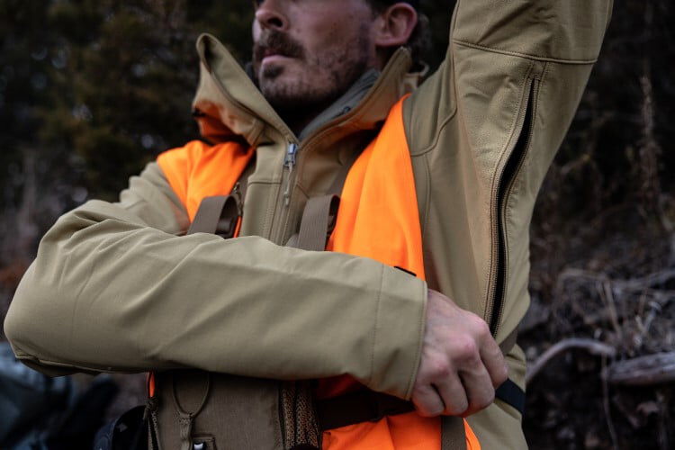 male wearing the De Havilland Jacket and an orange hunting vest while unzipping a pocket in the underarm