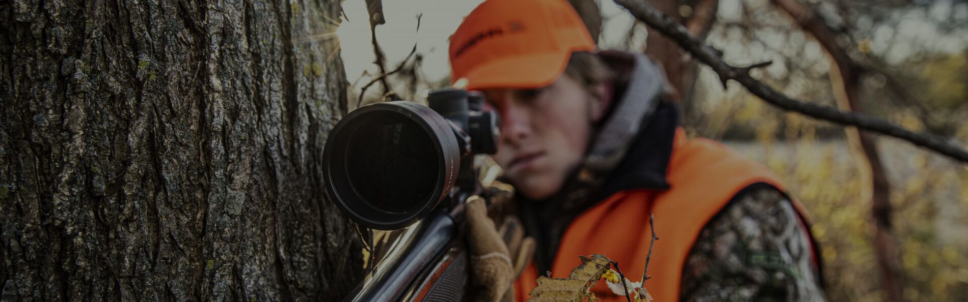 hunter looking down rifle scope while leaning against a tree
