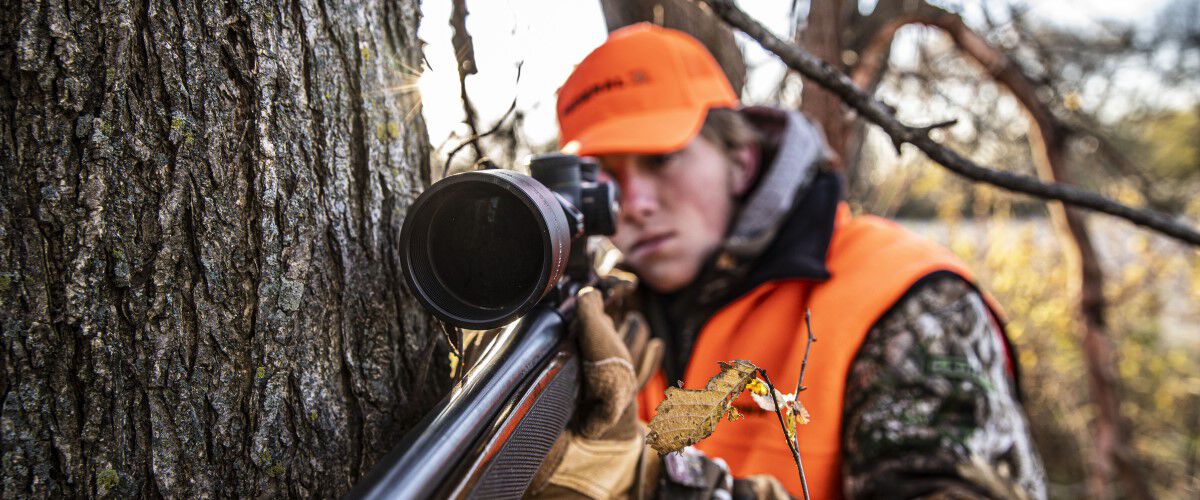 hunter looking down rifle scope while leaning against a tree