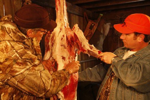 Two men removing the front quarter of a deer in a shed