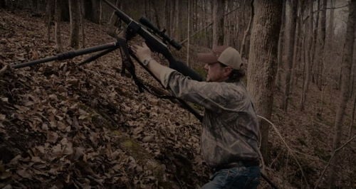 Jim Gilliland looking down a the scope of a rifle on a tripod angled up a hill